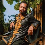 A fashionable man in a dashiki sits indoors surrounded by lush plants.