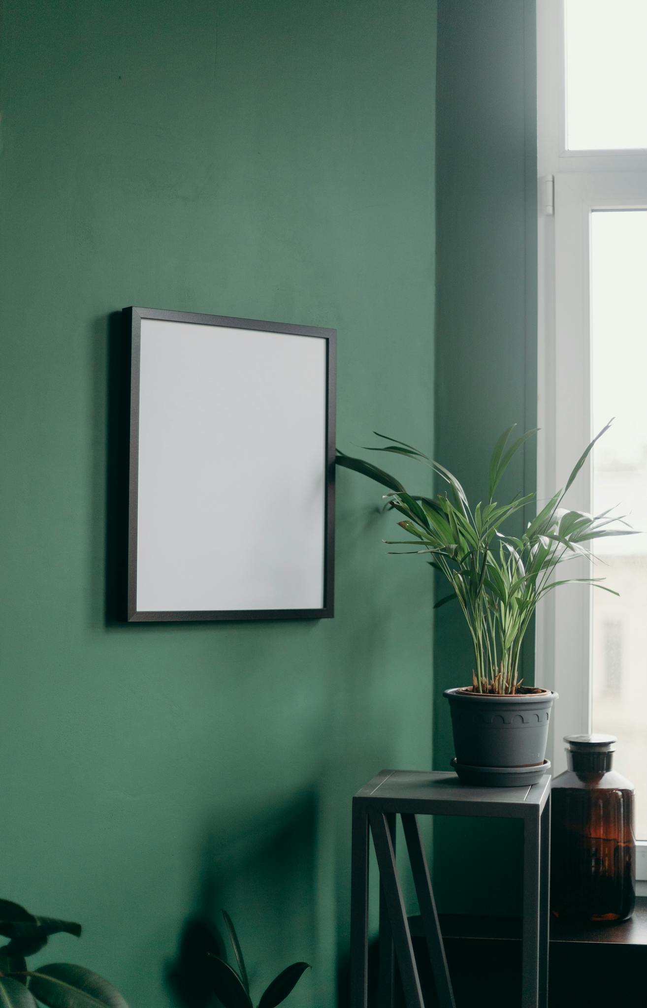 Stylish interior featuring a blank frame on a green wall and a potted plant by a window.