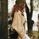 Stylish woman with curly hair in a trench coat and beanie, standing outdoors.